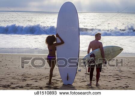 Stock Image - "Rear view of young surfing couple looking out from beach, Playa Del Rey, California, USA". Fotosearch