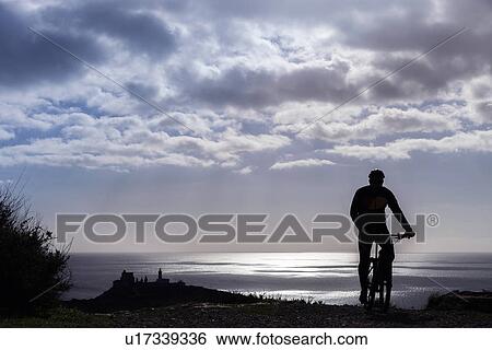 Stock Photograph - "Silhouetted rear view of male mountain biker on coastal path at dusk, Cagliari, Sardinia, Italy". Fotosearch
