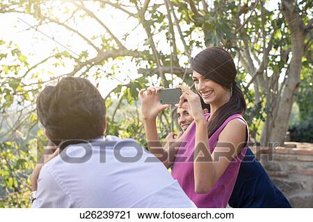 Stock Image - Three young adult friends photographing on smartphone in park. Fotosearch