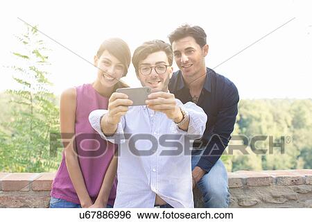 Three young adult friends taking smartphone selfie in front of wall View Large Photo Image Stock Photograph - Three young adult friends taking smartphone selfie in front of wall. Fotosearch