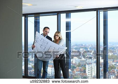 Stock Image - "Two architects looking at plans in front of office window with Brussels cityscape, Belgium". Fotosearch