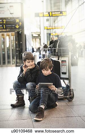 Two brothers sitting on airport luggage trolley looking at digital tablet View Large Photo Image Stock Photo - Two brothers sitting on airport luggage trolley looking at digital tablet. Fotosearch