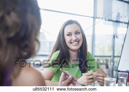 Stock Image - Two businesswomen gossiping at office desk over working lunch. Fotosearch