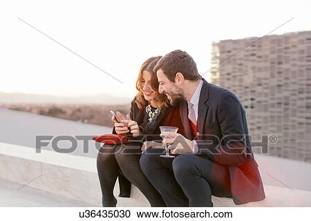 Stock Image - "Two friends drinking together at party, woman showing man her mobile phone". Fotosearch