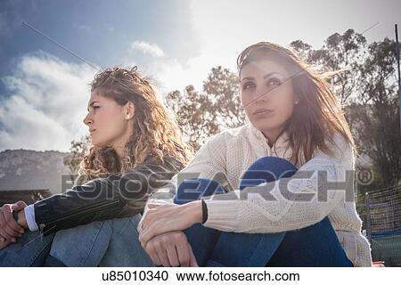 Stock Image - Two sullen young women friends sitting on breezy beach. Fotosearch
