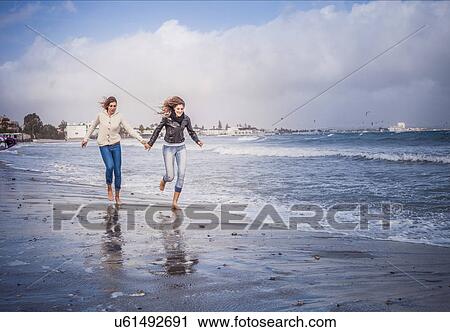 Stock Image - Two young women friends running hand in hand on beach. Fotosearch