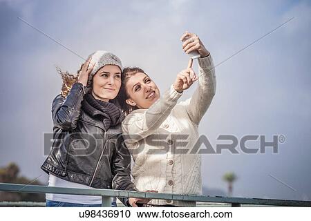 Stock Image - Two young women friends using touchscreen for smartphone selfie at coast. Fotosearch