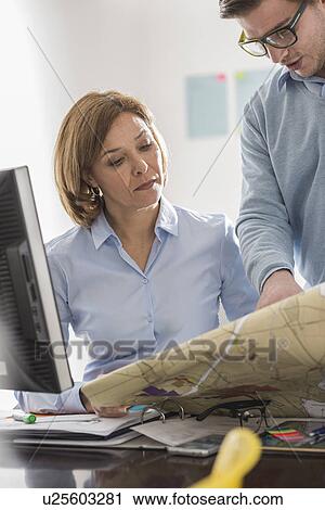 Stock Image - Young businessman looking at blueprint with architect at office desk. Fotosearch