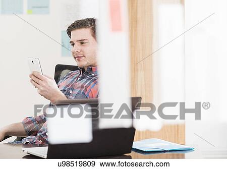 Young businessman looking at smartphone at office desk View Large Photo Image Stock Photo - Young businessman looking at smartphone at office desk. Fotosearch