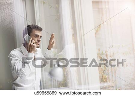 Stock Photo - Young businessman looking out of office window whilst chatting on smartphone. Fotosearch