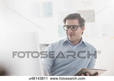 Stock Image - Young businessman using computer and digital tablet at office desk. Fotosearch