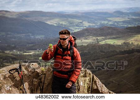 Stock Image - "Young male hiker taking smartphone selfie from mountain, The Lake District, Cumbria, UK". Fotosearch