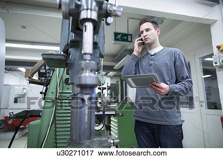 Stock Photo - Young male technician chatting on smartphone whilst using digital tablet in workshop. Fotosearch