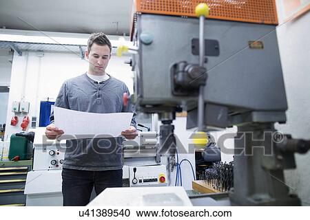 Stock Image - Young male technician looking at blueprint in workshop. Fotosearch