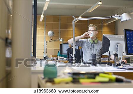 Stock Photo - Young man drinking coffee at office desk. Fotosearch