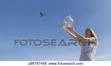 Stock Photo - Young woman releasing bird from cage toward blue sky. Fotosearch