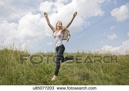 Stock Image - Young woman running through field with arms in air. Fotosearch