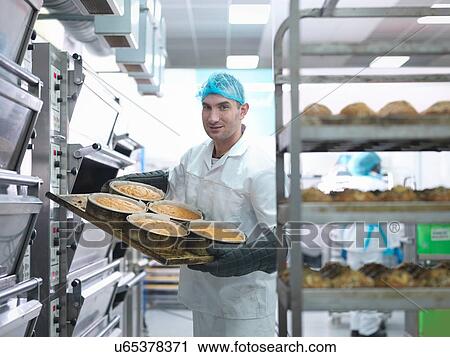 Stock Image - Baker taking cakes out of oven in cake factory. Fotosearch