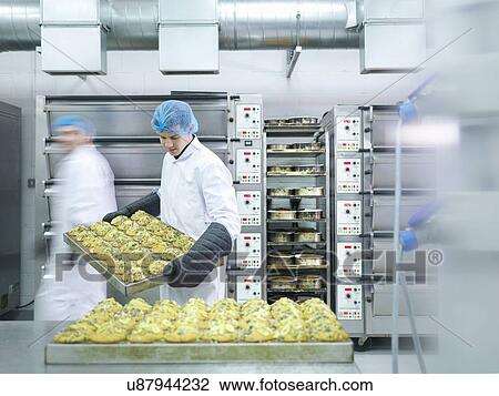 Stock Image - Baker taking cakes out of oven in cake factory. Fotosearch