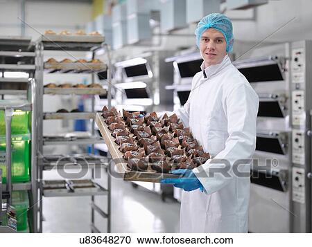 Stock Image - "Baker with tray of fresh cakes in cake factory, portrait". Fotosearch