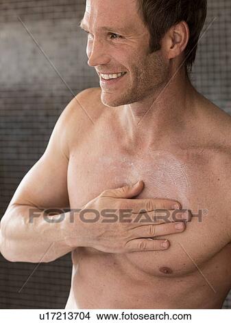 Picture - Bare chested muscular man in bathroom rubbing moisturizer on chest. Fotosearch