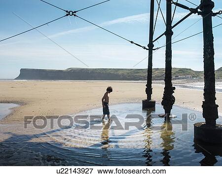 Stock Photo - Boy paddling in water under pier at beach. Fotosearch