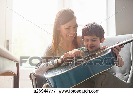 Stock Photo - Boy sitting on mother's lap in sunlit room and learning to play guitar. Fotosearch
