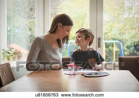 Stock Image - Boy smiling at mother as he paints on paper at table in living room. Fotosearch