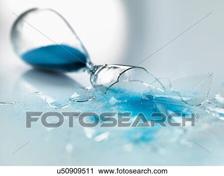 Stock Image - Broken glass hourglass with blue sand pouring out. Fotosearch