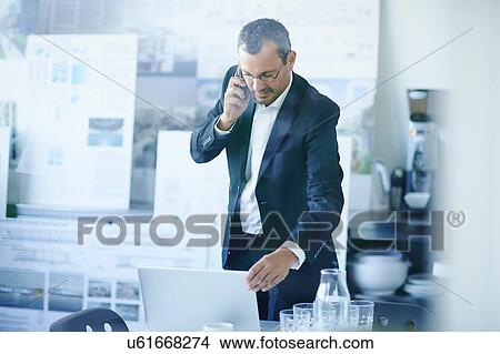 Business man chatting on smartphone whilst using office laptop View Large Photo Image Picture - Business man chatting on smartphone whilst using office laptop. Fotosearch