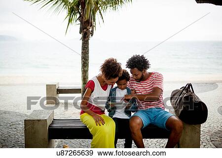 Stock Photo - "Couple and son sitting on park bench using digital tablet, Ipanema beach, Rio De Janeiro, Brazil". Fotosearch