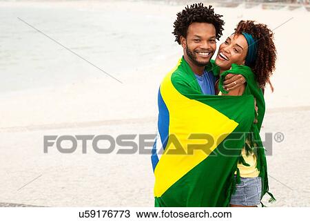 Stock Image - "Couple hugging wrapped in Brazilian flag on Ipanema beach, Rio De Janeiro, Brazil". Fotosearch