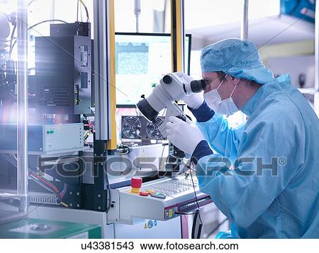 Stock Image - Electronics worker checking component with microscope in clean room. Fotosearch