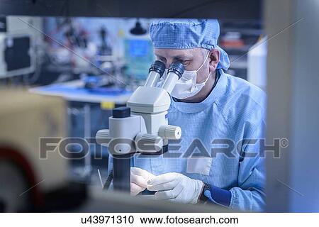 Stock Image - Electronics worker checking component with microscope in clean room. Fotosearch