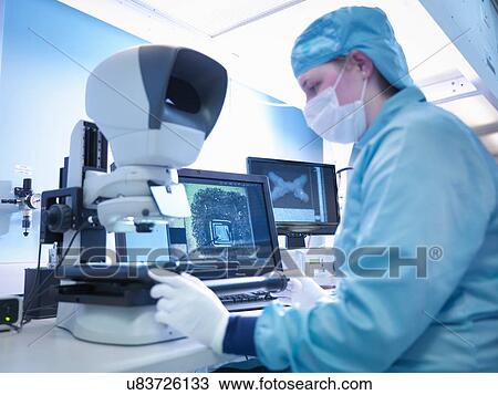 Stock Image - Electronics worker checking components with microscope in clean room. Fotosearch