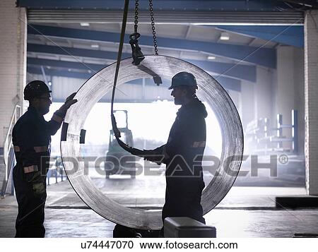 Stock Image - Engineers using crane to lift large steel tube in engineering factory. Fotosearch