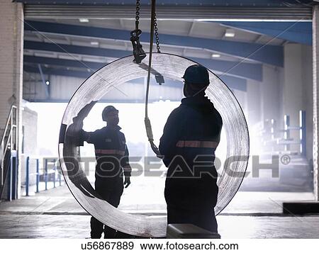 Stock Photo - Engineers using crane to lift large steel tube in engineering factory. Fotosearch
