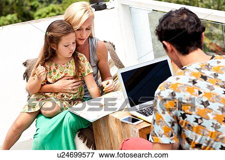 "Family sitting at table on balcony, mother and daughter looking through book while father uses laptop" View Large Photo Image Stock Photo - "Family sitting at table on balcony, mother and daughter looking through book while father uses laptop". Fotosearch