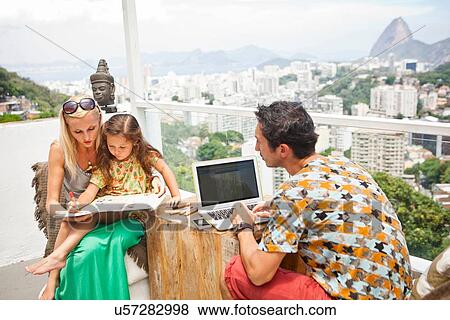 Stock Photo - "Family sitting at table on balcony, mother and daughter looking through book while father uses laptop". Fotosearch
