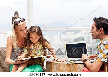 "Family sitting at table on balcony, mother and daughter looking through book while father uses laptop" View Large Photo Image Stock Photo - "Family sitting at table on balcony, mother and daughter looking through book while father uses laptop". Fotosearch