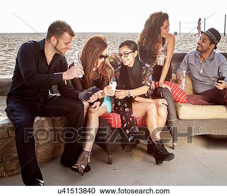 Stock Image - Group of friends at a beach bar talking and looking at smart phone. Fotosearch