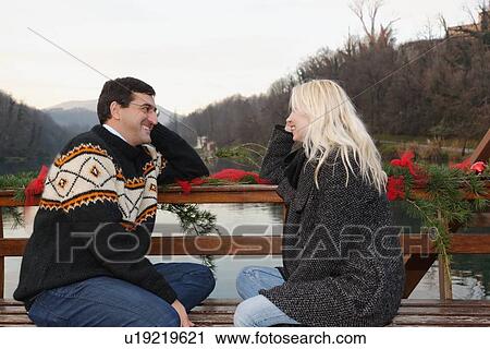 Stock Image - "Heterosexual couple sitting face to face on bench, beside lake, Lombardy, Italy". Fotosearch