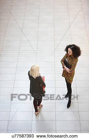 Stock Image - High angle view of two businesswomen chatting in office atrium. Fotosearch