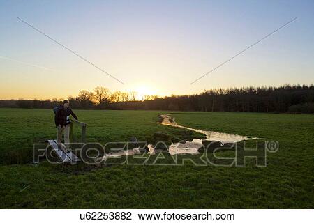 Stock Image - Male hiker crossing footbridge in field at sunset. Fotosearch