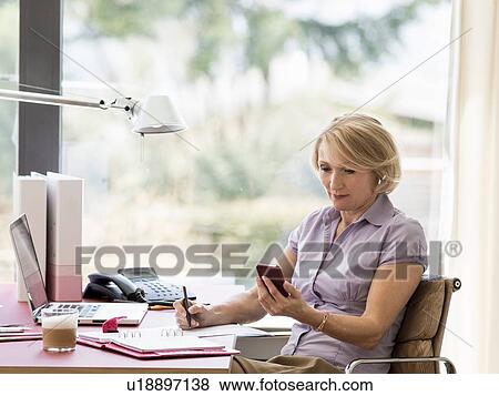 Stock Photo - Mature businesswoman reading text message on smartphone at home desk. Fotosearch