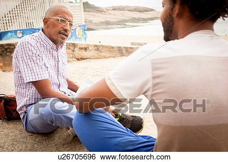 Stock Photograph - "Mature man sitting on Ipanema beach chatting to son, Rio De Janeiro, Brazil". Fotosearch