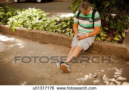 Stock Photograph - "Mature man sitting on sidewalk texting on smartphone, Rio De Janeiro, Brazil". Fotosearch