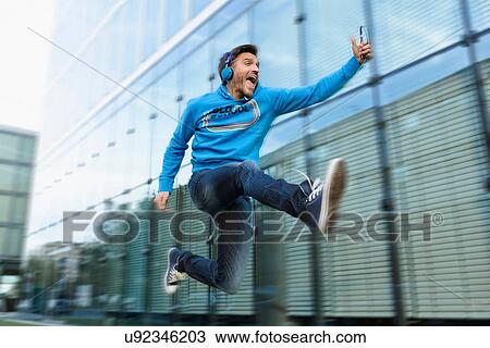 Stock Image - Mature man with headphones and smartphone jumping mid air on street. Fotosearch