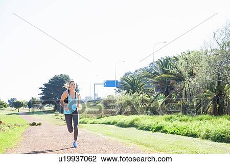 Stock Image - Mid adult woman running on dirt track in park. Fotosearch