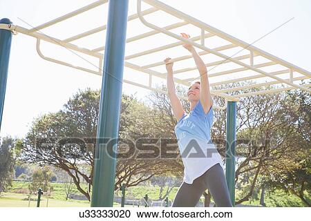 Stock Image - Mid adult woman training in park on monkey bars. Fotosearch